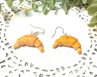 Pode incluir: Um par de brincos em forma de croissant com ganchos prateados. Os croissants são de cor castanho claro.