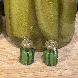 May include: A pair of novelty earrings shaped like miniature pickle jars. The earrings are green with vertical stripes, gold lids, and silver-colored hooks. A large jar of pickles is in the background, sitting on a wooden surface.