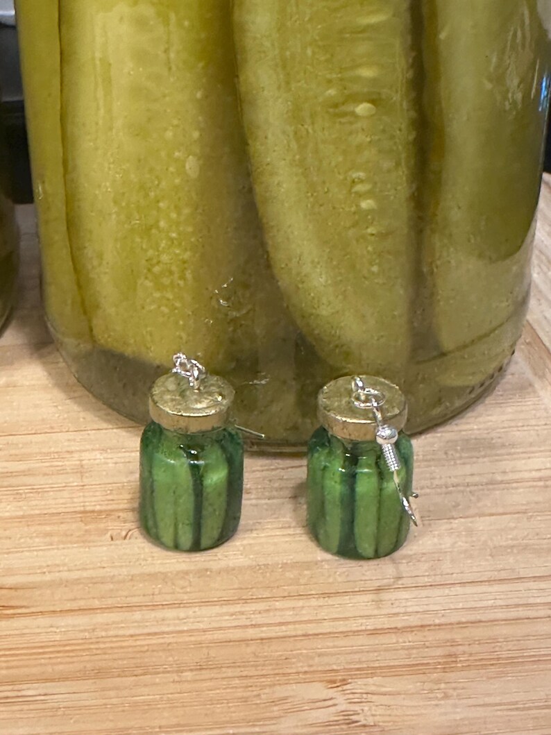 May include: A pair of novelty earrings shaped like miniature pickle jars. The earrings are green with vertical stripes, gold lids, and silver-colored hooks. A large jar of pickles is in the background, sitting on a wooden surface.