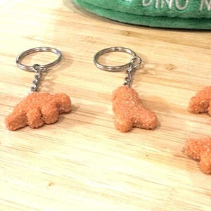 May include: Orange dinosaur claw keyrings with silver key rings. The keyrings are made of a textured material and are arranged on a wooden surface. A green plush item is visible in the background.