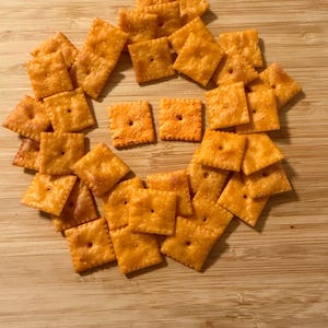 Puede incluir: Una colecci&oacute;n de galletas de queso cuadradas y naranjas dispuestas sobre una superficie de madera de color marr&oacute;n claro. Las galletas tienen una superficie ligeramente texturizada y un peque&ntilde;o agujero en el centro. La disposici&oacute;n forma una forma circular.