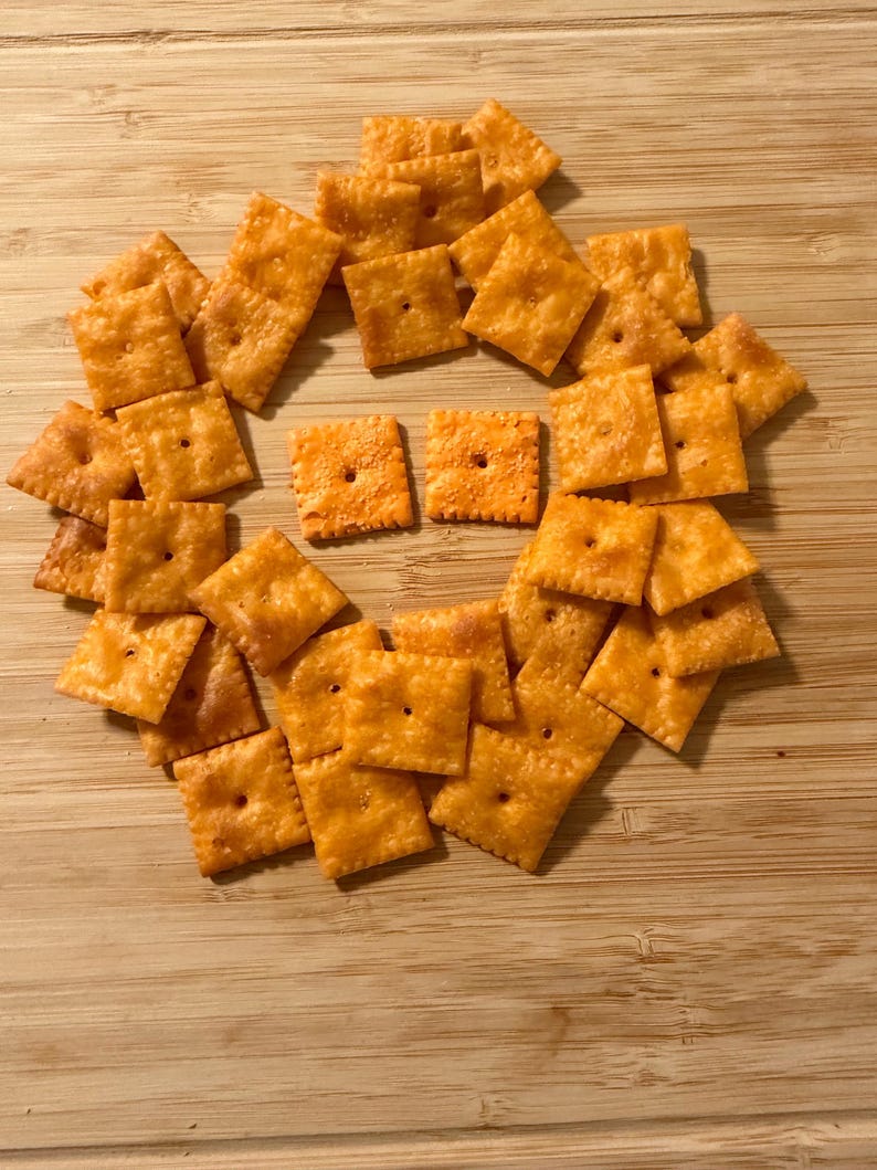 Puede incluir: Una colecci&oacute;n de galletas de queso cuadradas y naranjas dispuestas sobre una superficie de madera de color marr&oacute;n claro. Las galletas tienen una superficie ligeramente texturizada y un peque&ntilde;o agujero en el centro. La disposici&oacute;n forma una forma circular.