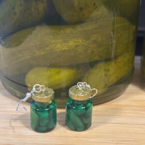 May include: A pair of novelty earrings shaped like miniature jars of pickles. The earrings are made of green resin with tiny pickle shapes inside. The jar lids are gold-colored, and the earrings have silver-colored hooks.