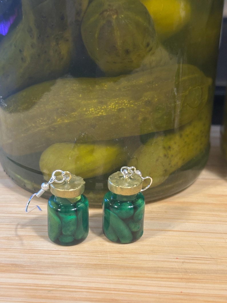 May include: A pair of novelty earrings shaped like miniature jars of pickles. The earrings are made of green resin with tiny pickle shapes inside. The jar lids are gold-colored, and the earrings have silver-colored hooks.