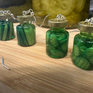 May include: Novelty earrings featuring miniature glass jars filled with green pickle slices. The jars have gold-colored lids and silver earring hooks. The earrings are displayed on a wooden surface, with larger jars of pickles in the background.