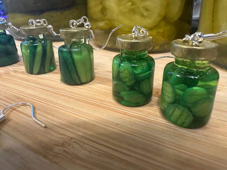 May include: Novelty earrings featuring miniature glass jars filled with green pickle slices. The jars have gold-colored lids and silver earring hooks. The earrings are displayed on a wooden surface, with larger jars of pickles in the background.