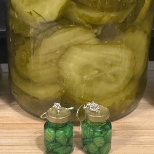 May include: A pair of novelty earrings shaped like miniature pickle jars, filled with green pickle slices. The earrings have silver-colored hooks and gold-colored lids. A large jar of sliced pickles is in the background.