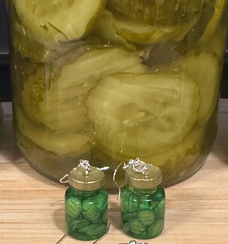 May include: A pair of novelty earrings shaped like miniature pickle jars, filled with green pickle slices. The earrings have silver-colored hooks and gold-colored lids. A large jar of sliced pickles is in the background.