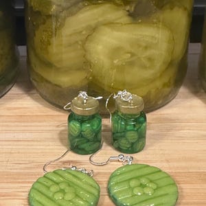 May include: A pair of novelty earrings featuring a pickle jar design. The earrings have a miniature jar filled with green pickles, and a matching green pickle slice. The earrings are on a wooden surface.
