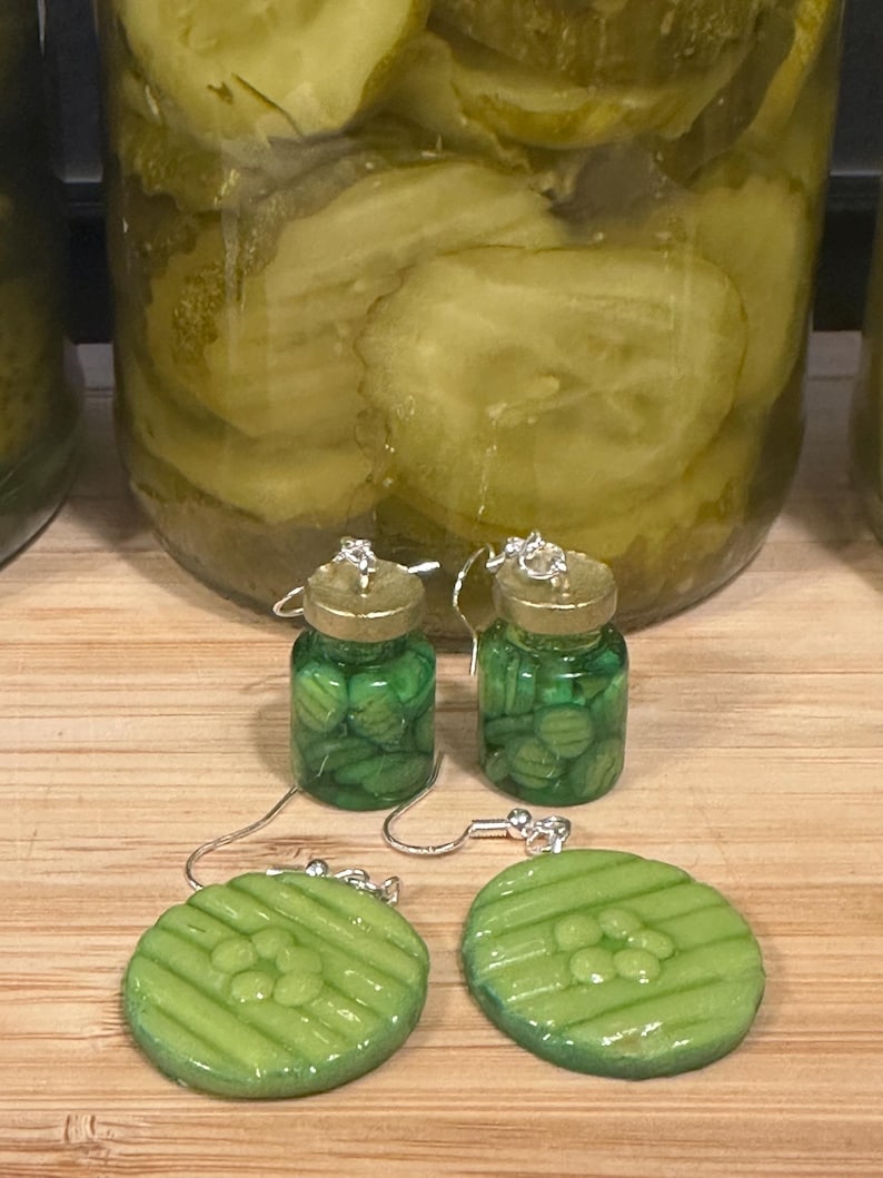 May include: A pair of novelty earrings featuring a pickle jar design. The earrings have a miniature jar filled with green pickles, and a matching green pickle slice. The earrings are on a wooden surface.