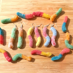 Puede incluir: Una colección de caramelos de gominola con forma de gusano de colores y un par de pendientes de gominola con forma de gusano. Los pendientes tienen un gancho plateado y están diseñados para parecerse a los caramelos. Los caramelos son de varios colores, como rojo, amarillo, verde y azul, y están colocados sobre una superficie de madera.