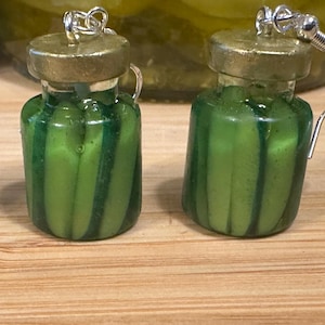 May include: A pair of novelty earrings shaped like miniature pickle jars. The earrings are green with vertical stripes, topped with a gold-colored lid and silver ear hooks. The background shows a jar of pickles on a wooden surface.