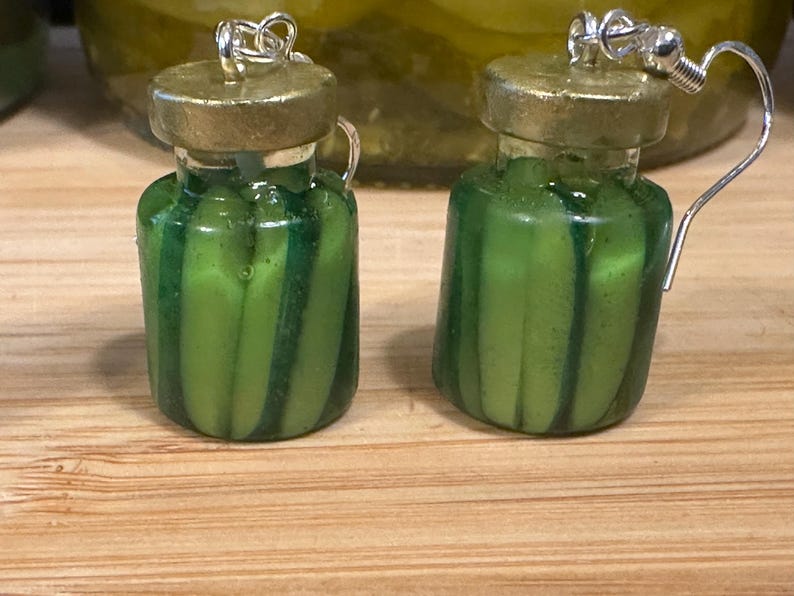 May include: A pair of novelty earrings shaped like miniature pickle jars. The earrings are green with vertical stripes, topped with a gold-colored lid and silver ear hooks. The background shows a jar of pickles on a wooden surface.