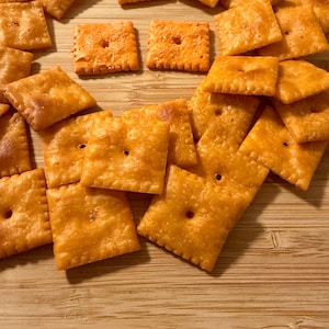 Puede incluir: Un conjunto de galletas saladas cuadradas, de color dorado y ligeramente texturizadas, cada una con un agujero central. Las galletas est&aacute;n dispuestas sobre una superficie de madera clara. La imagen muestra un aperitivo, con su color y forma distintivos.