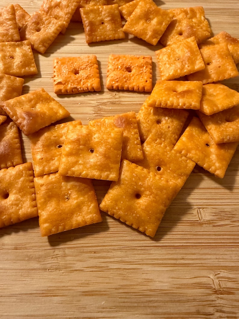 Puede incluir: Un conjunto de galletas saladas cuadradas, de color dorado y ligeramente texturizadas, cada una con un agujero central. Las galletas est&aacute;n dispuestas sobre una superficie de madera clara. La imagen muestra un aperitivo, con su color y forma distintivos.