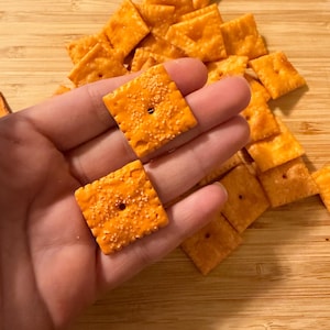 Puede incluir: Primer plano de galletas cuadradas naranjas con una superficie texturizada y un peque&ntilde;o agujero en el centro. Las galletas est&aacute;n espolvoreadas con condimentos. Un mont&oacute;n de galletas en el fondo.