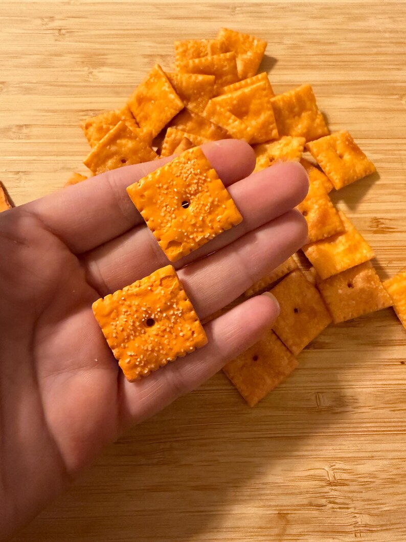 Puede incluir: Primer plano de galletas cuadradas naranjas con una superficie texturizada y un peque&ntilde;o agujero en el centro. Las galletas est&aacute;n espolvoreadas con condimentos. Un mont&oacute;n de galletas en el fondo.