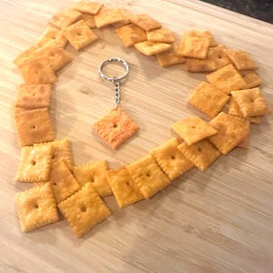 May include: A heart shape made of golden-brown crackers surrounds a keychain with a single cracker charm. The crackers are arranged on a light-colored wooden cutting board, creating a playful and unique design.