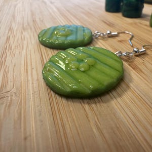 May include: A pair of novelty earrings featuring green, pickle-shaped pendants with silver-colored fish hook ear wires. The pickle slices have a glossy finish and are detailed with ridges and small, round embellishments.