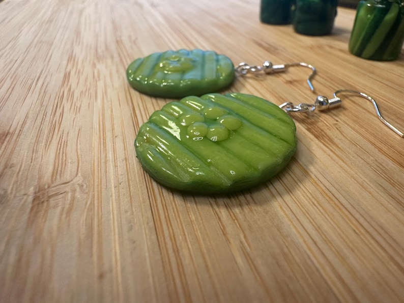 May include: A pair of novelty earrings featuring green, pickle-shaped pendants with silver-colored fish hook ear wires. The pickle slices have a glossy finish and are detailed with ridges and small, round embellishments.