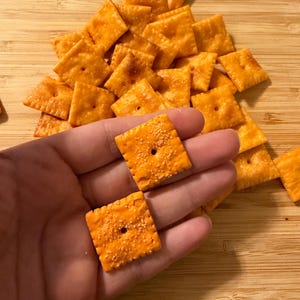 Puede incluir: Primer plano de galletas cuadradas naranjas con un agujero en el centro y un borde texturizado. Las galletas est&aacute;n esparcidas sobre una superficie de madera, con dos galletas en una mano.