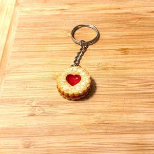 Pode incluir: Um porta-chaves com uma réplica em miniatura e detalhada de um biscoito sanduíche com geleia em forma de coração. O biscoito tem um exterior castanho-dourado, um recheio de geleia vermelha e um recorte em forma de coração. O porta-chaves é preso a um anel prateado.