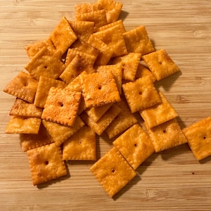 Puede incluir: Un mont&oacute;n de galletas cuadradas y anaranjadas, cada una con un agujero central. Est&aacute;n dispuestas sobre una tabla de madera marr&oacute;n claro. Las galletas tienen una superficie ligeramente texturizada.