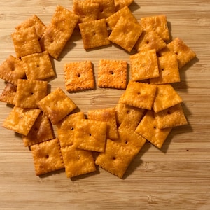 Puede incluir: Un conjunto de galletas cuadradas de queso naranja dispuestas sobre una superficie de madera. Cada galleta tiene un peque&ntilde;o agujero en el centro. Las galletas son de color naranja dorado, lo que sugiere que est&aacute;n horneadas.