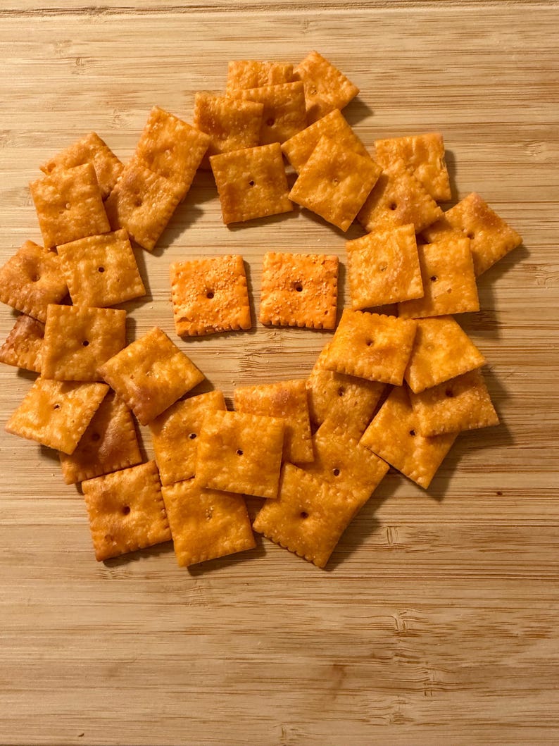 Puede incluir: Un conjunto de galletas cuadradas de queso naranja dispuestas sobre una superficie de madera. Cada galleta tiene un peque&ntilde;o agujero en el centro. Las galletas son de color naranja dorado, lo que sugiere que est&aacute;n horneadas.