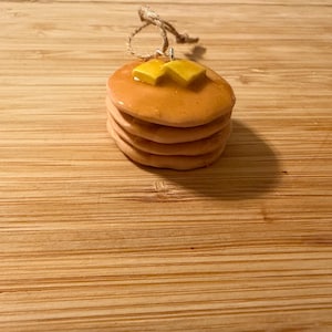 May include: A stack of four pancakes with two square pats of butter on top. The pancakes are light brown and appear to be drizzled with syrup. A twine loop is attached to the top. The background is a wooden surface.