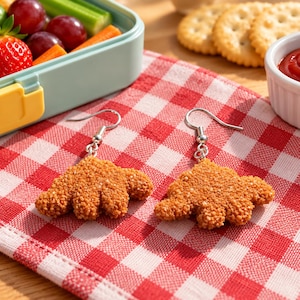 Könnte beinhalten: Ein Paar Ohrhänger in Form von panierten Hähnchen-Nuggets. Jeder Ohrring hat eine texturierte, goldbraune, knusprig aussehende Oberfläche, die an einem silberfarbenen Metallhaken befestigt ist. Die Ohrringe liegen auf einem rot-weiß karierten Tuch.