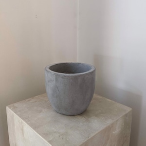 May include: A grey concrete planter with a rounded shape. The planter is sitting on a grey concrete pedestal.