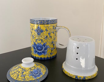 Puede incluir: Una taza de té amarilla y azul con un diseño floral y una tapa e infusor a juego. La taza de té tiene un asa y el infusor tiene agujeros para que las hojas de té se infusen.