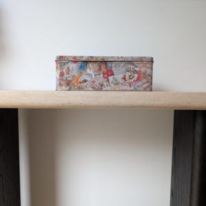 May include: A rectangular tin box with a colorful collage design featuring various images and patterns. The box is placed on a light-colored shelf against a white wall, with a black frame above.