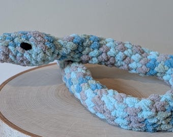 May include: A crocheted snake toy made of blue, grey, and green yarn. The snake is curled up in a circle and has a small, black eye.