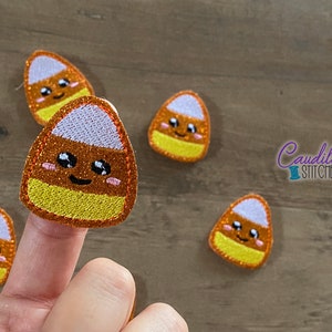 Op de afbeelding: Geborduurde candy corn patches met glinsterende oranje, gele en witte secties. Elke patch heeft een lachend gezicht met roze wangen en zwarte ogen. De patches zijn verspreid over een houten oppervlak. De tekst "Caudill Stitches" is zichtbaar.