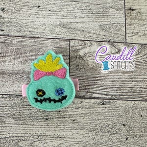 May include: A green and blue felt character with a yellow crown and pink bow. The character has a stitched face with black eyes and a stitched mouth.