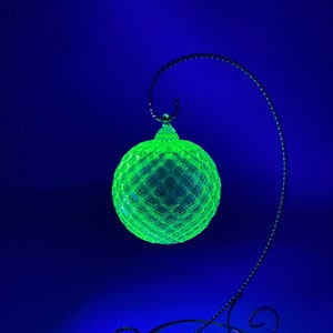 Puede incluir: Un adorno navideño de vidrio verde con un patrón de diamante cuelga de un soporte de metal negro sobre un fondo azul.