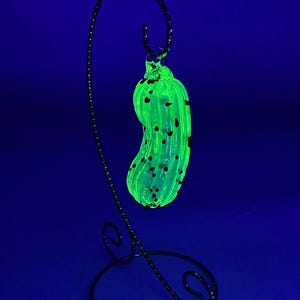 Puede incluir: Un adorno de vidrio verde en forma de pepinillo con motas negras que cuelga de un soporte de metal negro. El adorno brilla bajo una luz negra.
