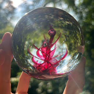 May include: A clear glass sphere with a vibrant, abstract red design inside. The design features flowing, organic shapes. The sphere is held up against a backdrop of green foliage and sunlight, creating a visually striking effect.
