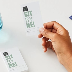 May include: White business cards with the text "SIT BY ME!" in green, a logo, and a contact line. A stack of cards and a single card are visible. A glass of blue liquid and a blue pen are also in the image.