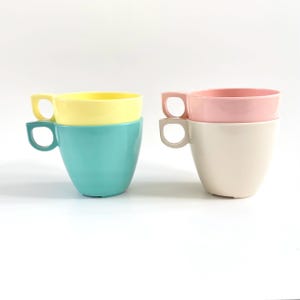 May include: Four colourful cups stacked in pairs. One pair features a yellow cup stacked on a teal cup. The other pair has a pink cup on top of a cream-coloured cup. Each cup has a unique handle.