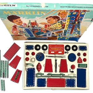 Puede incluir: Juego de construcción Marklin vintage con una caja con ilustraciones de niños. El juego incluye varias piezas metálicas en rojo, azul y verde, junto con ruedas y otros componentes. La caja es azul claro y blanca.