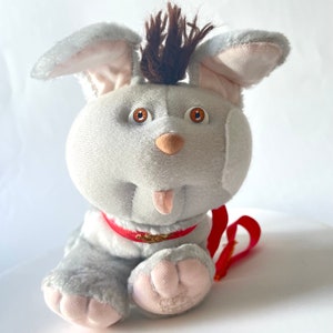 May include: A grey plush toy dog with brown fur on its head, brown eyes, and a red collar. The dog has its tongue sticking out and is wearing a red lead.