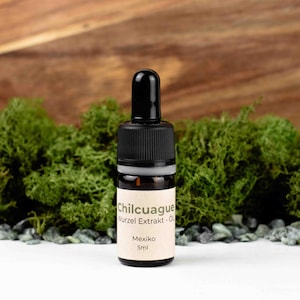 Puede incluir: Una pequeña botella de vidrio con un tapón cuentagotas negro, etiquetada como "Chilcuague Wurzel Extrakt - Öl" y "Mexiko 5ml." La botella contiene un líquido transparente. El fondo presenta musgo verde y una superficie de madera.