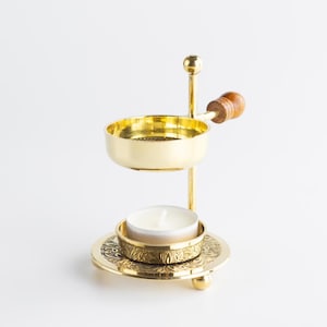Puede incluir: Un quemador de aceite de aromaterapia de metal dorado con un mango de madera. El quemador tiene una base redonda con patrones florales, un plato pequeño para una vela de té y un cuenco encima para aceites esenciales. Una vela blanca encendida está en la base.