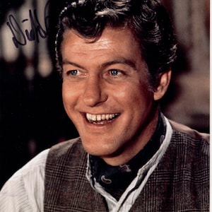 May include: A color photograph of a smiling man with blue eyes and dark hair. He wears a white striped shirt, a brown plaid vest, and a patterned scarf. The image includes an autograph in the upper left corner and a certification sticker in the lower right.