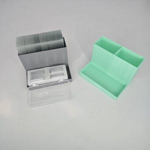 May include: Two storage containers. One is gray with a clear lid and contains small, clear, rectangular items. The other is mint green and has two compartments. The containers are on a white surface.