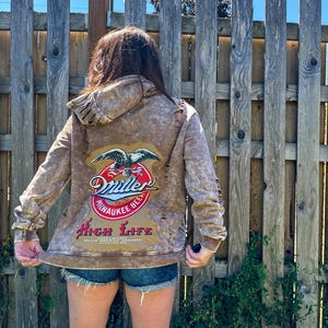 Könnte beinhalten: Ein brauner Kapuzenpullover mit dem Miller High Life Bierlogo auf dem Rücken. Das Logo zeigt einen Adler, die Worte "Miller", "Milwaukee Beer" und "High Life". Der Kapuzenpullover hat einen abgenutzten, Vintage-Look.