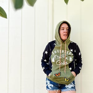 Puede incluir: Una sudadera con capucha verde oliva y negra con el texto "Adventure Calls" y un gráfico de montaña. Las mangas son negras con estrellas blancas. Dos parches de mariposa están en el bolsillo delantero. La persona lleva pantalones cortos vaqueros.